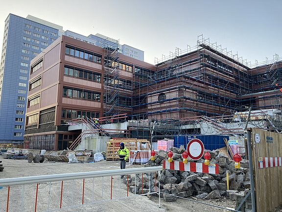 Blick auf die Baustelle und Fassade des Schulgebäudes und dahinter ein hohes Wohngebäude
