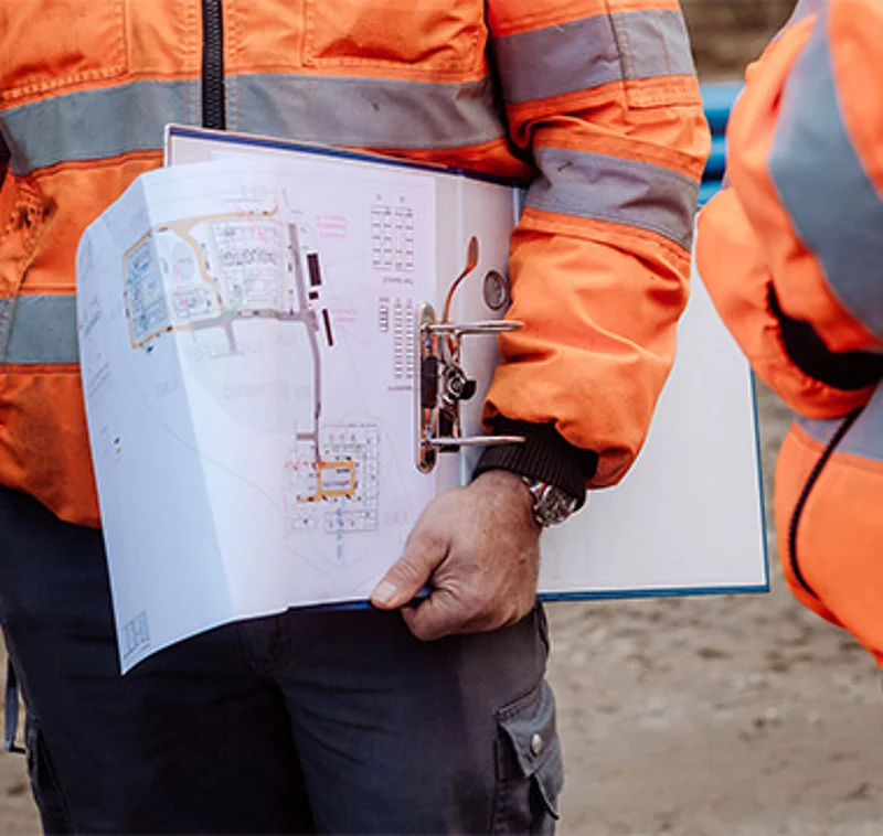 Close-up photograph of civil engineers on a construction site. Under their arm, they are carrying a project folder containing construction plans. 