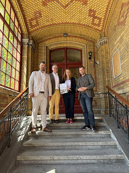 Mitarbeiter HOWOGE und Schulleiter mit Projektübersicht in der Hand in dem Eingang des Backsteingebäudes