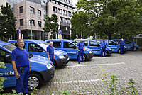 Sechs blaue Servicemobile der HOWOGE mit sechs Mitarbeitern in Blaumännern vor jeweils einem Mobil 