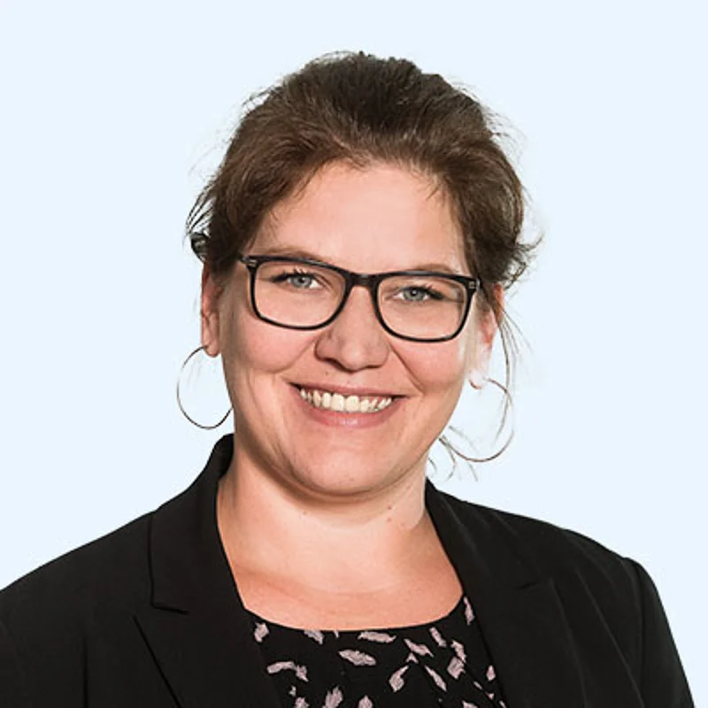 Eine Frau mittleren Alters mit lockigem, langen, braunem Haar und Brille. Sie lacht in die Kamera 