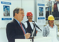 Eine Person hält eine Ansprache vor einem Podest, im Hintergrund erkennt man Bauarbeiter