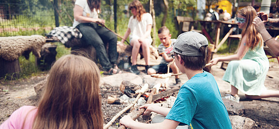 Kinder sitzen am Lagerfeuer und machen Stockbrot.