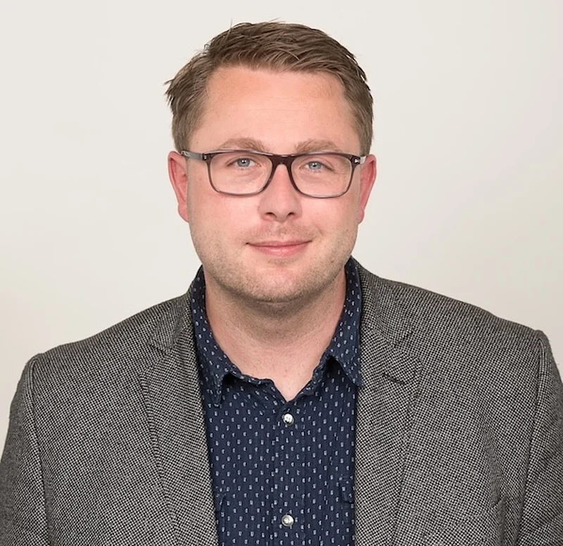 Ein Mann mittlerem Alters mit Brille und grauem Sakko. Lacht in Kamera