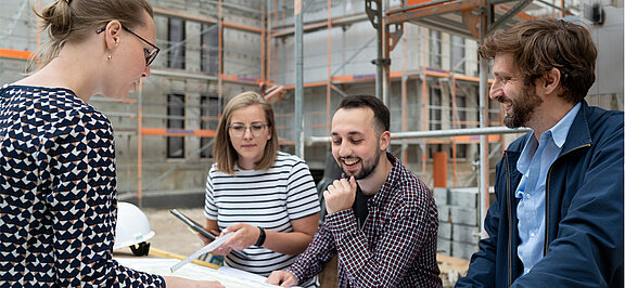 4 Azubis stehen vor einem Bauprojekt und besprechen sich.