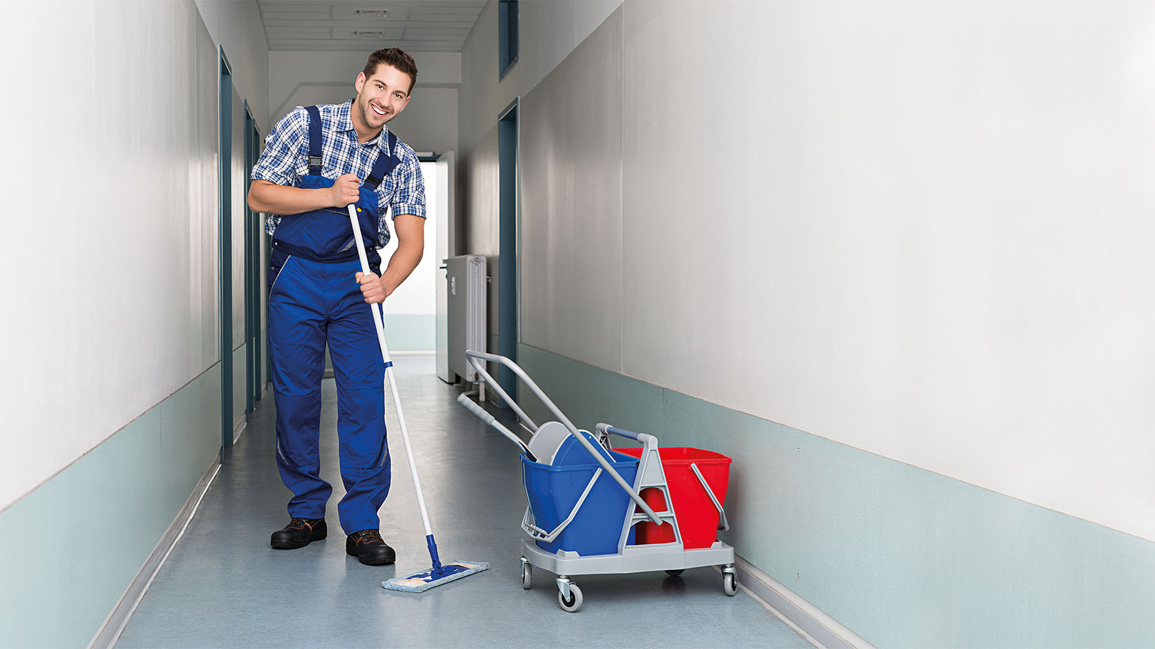 Foto einer lächelnden Reinigungskraft, die den Flur in einem Gebäude wischt. 