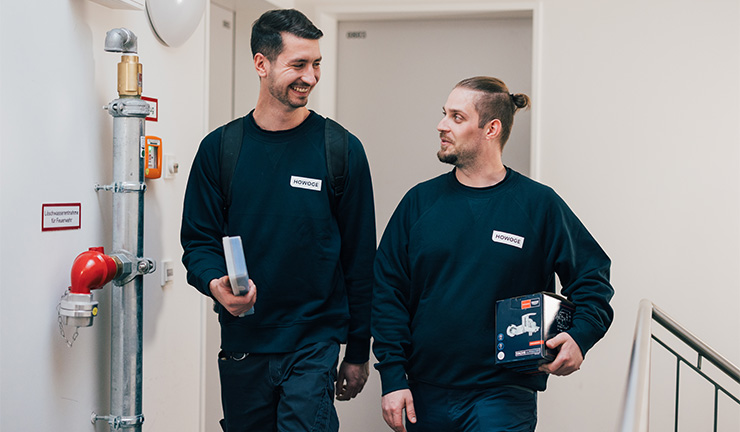 Zwei MItarbeiter der ATOZ Facility Solutions GmbH laufen einen Flur entlang und unterhalten sich. Dabei schauen sie sich an und lächeln.