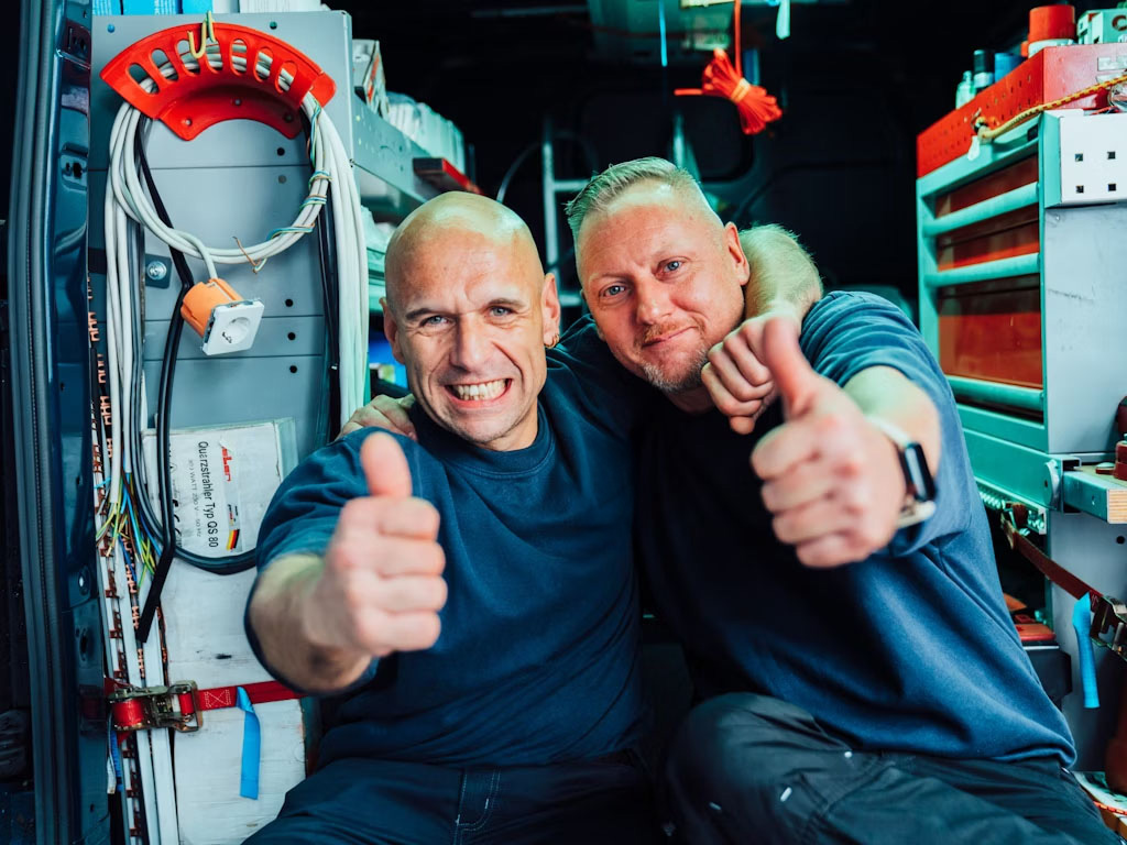 Zwei Elektriker lächeln und zeigen den Daumen nach oben in die Kamera.